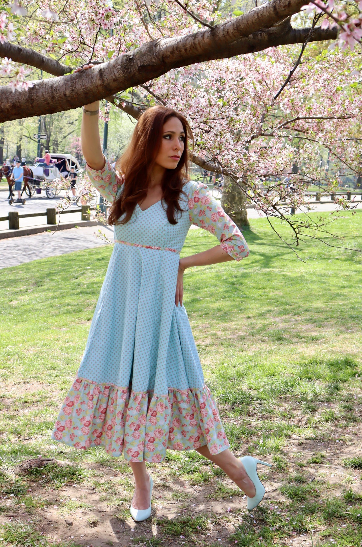 Model in aqua blue print midi dress with sleeves and lower ruffle in print and blue rose print holding out skirt and standing holding onto a tree limb.