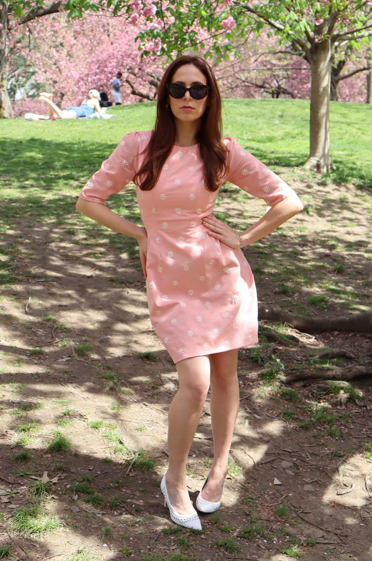 Model in peachy pink short dress with white daisy appliques looking forward in front of a cherry tree.