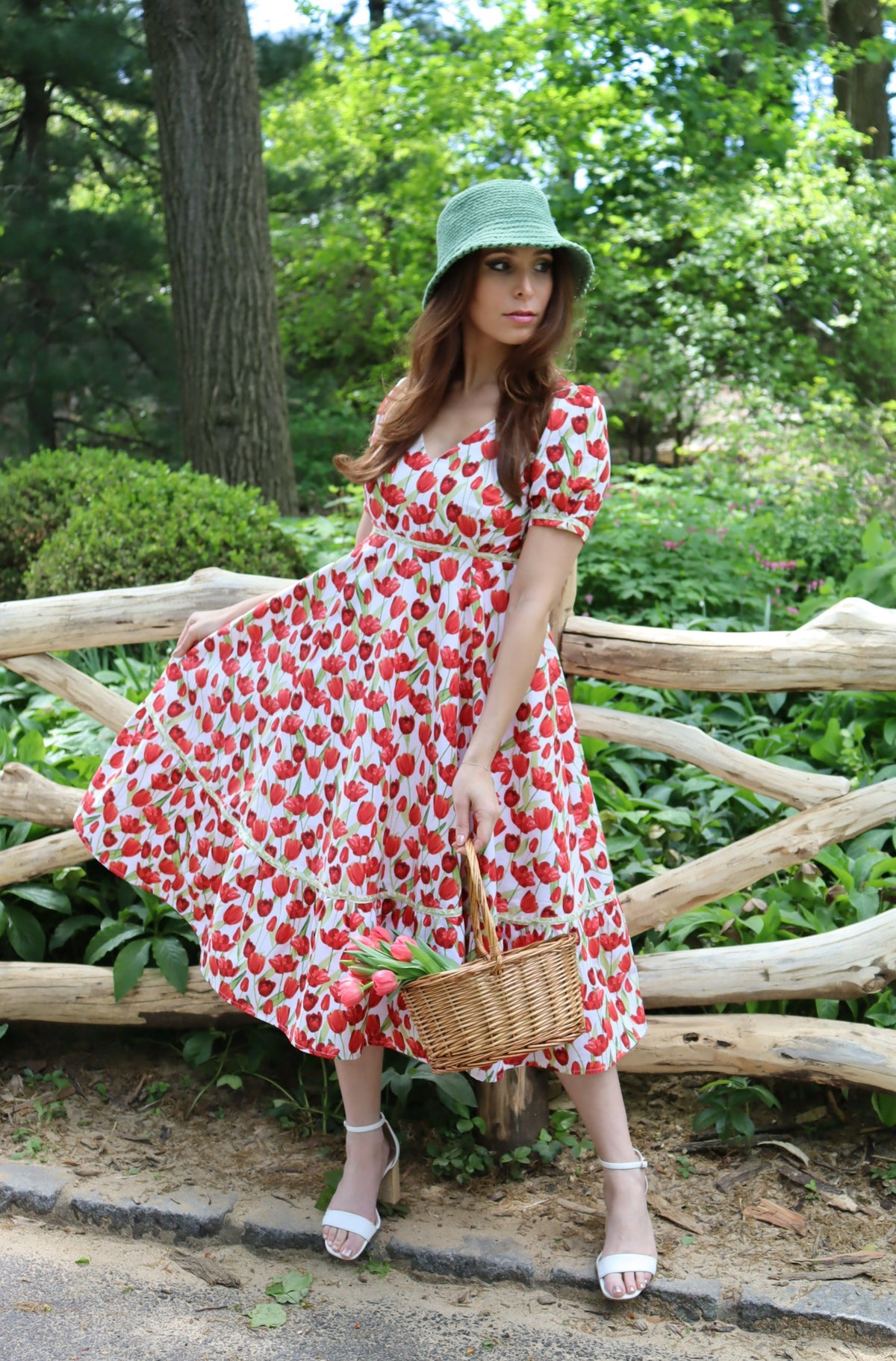 Model wearing midi length dress with a tulip print on white with a green bucket hat and holding a basket of tulips in front of a fence.