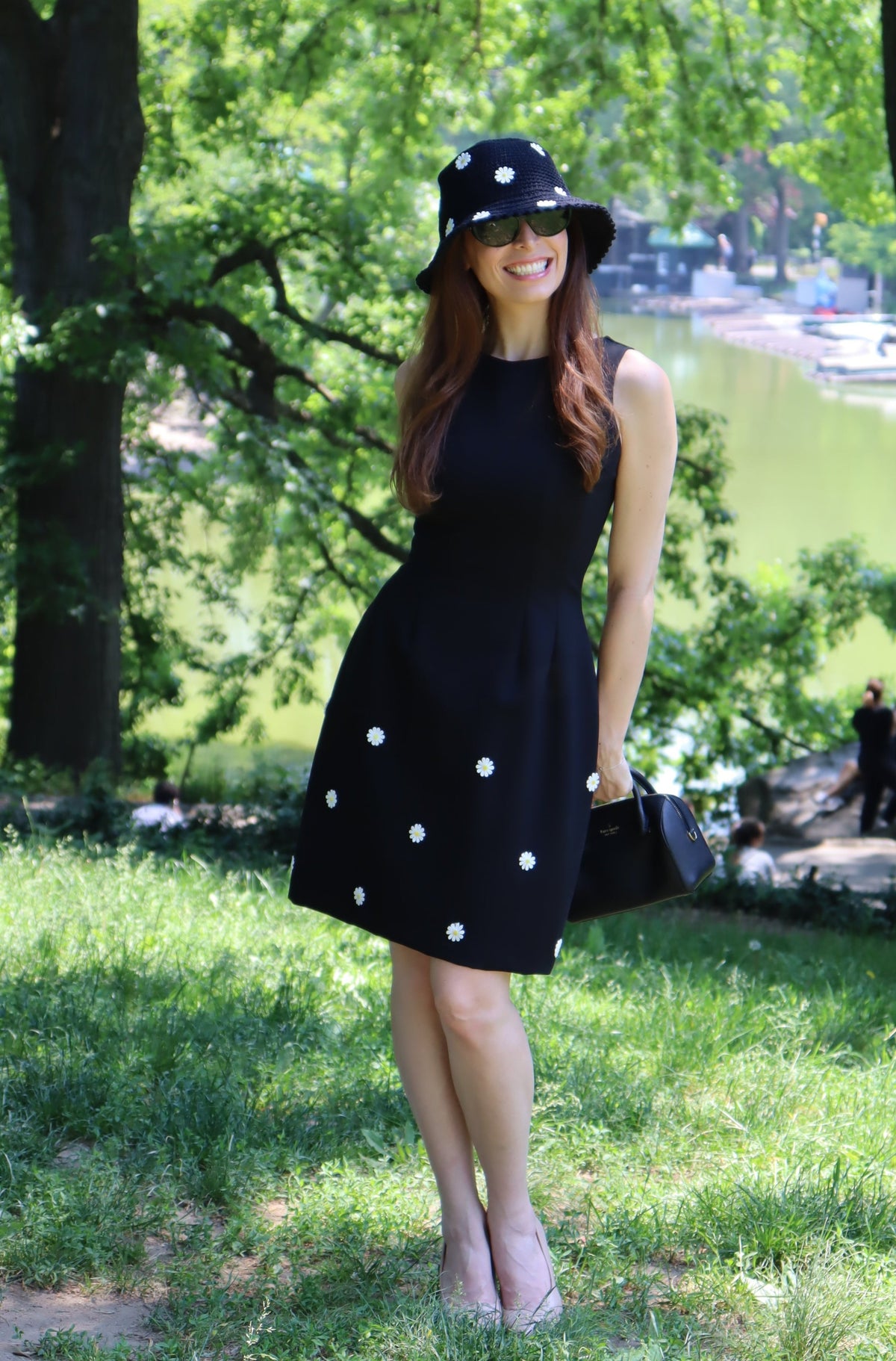 Model in a black dress with white daisy appliques standing in front of a tree and a lake.