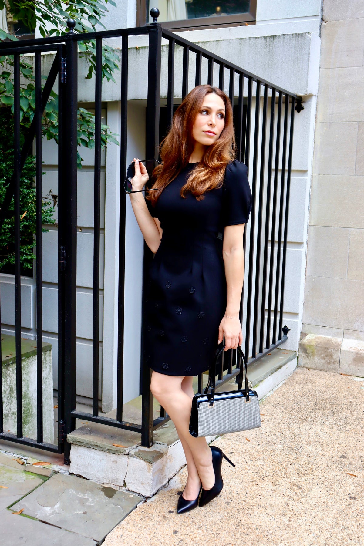 Model leaning on a fence wearing our Fall in Love Floral Appliquéd Black Ponte Dress, holding her sunglasses and a classic bag and heel combo