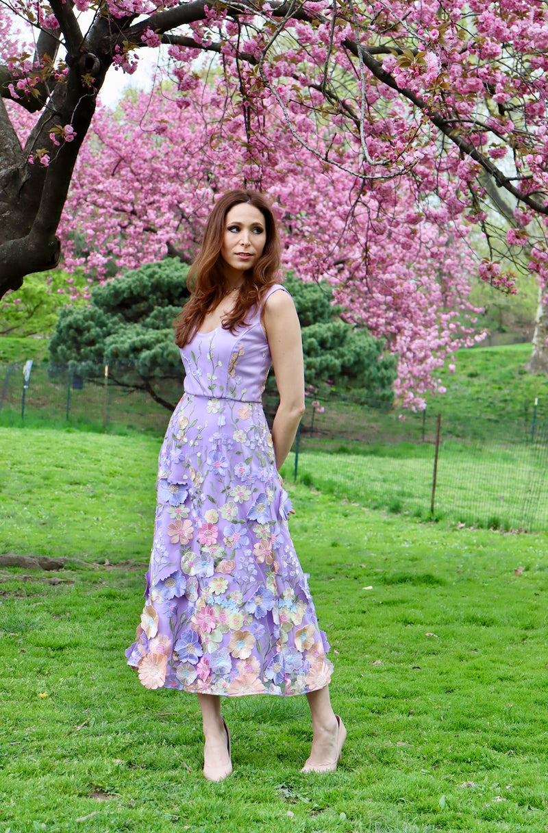 Model standing tall in lilac midi length sleeveless dress with satin appliquéd details.