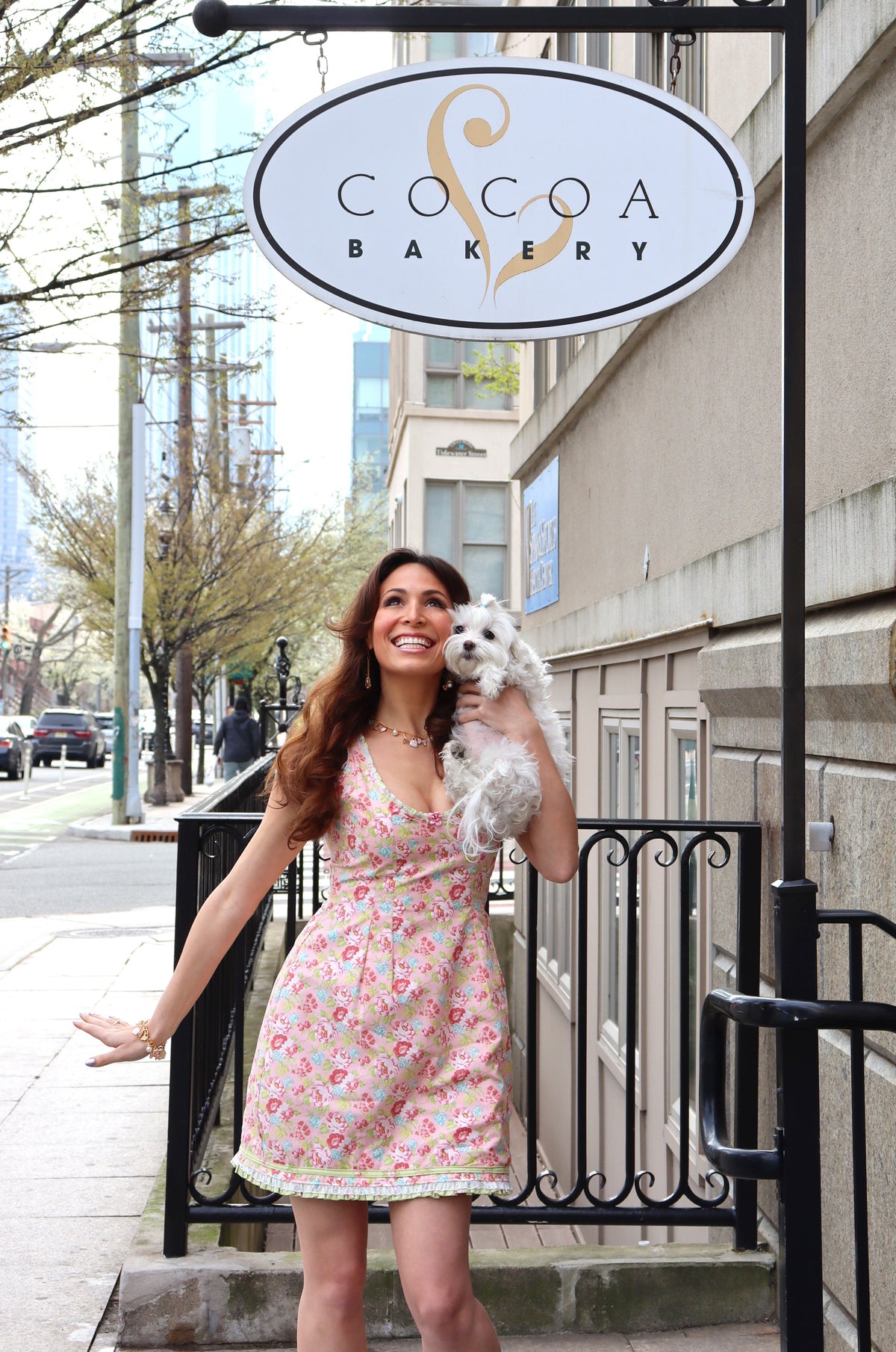 Model wearing a pink, green and blue floral print dress holding a white dog smiling at the sky.