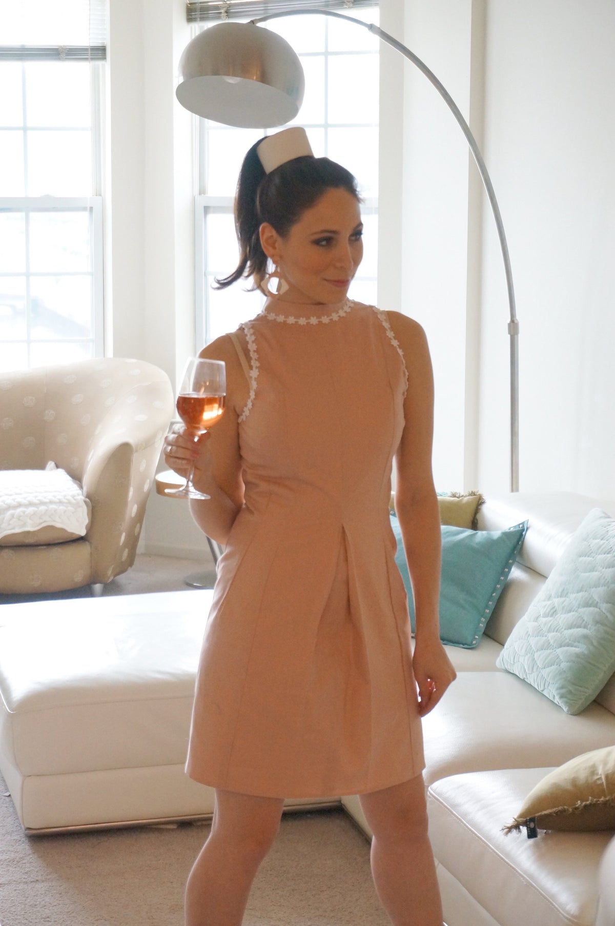 Model is wearing a pink dress with lace daisy trim and a front kick pleat looking off to the side in front of a couch.