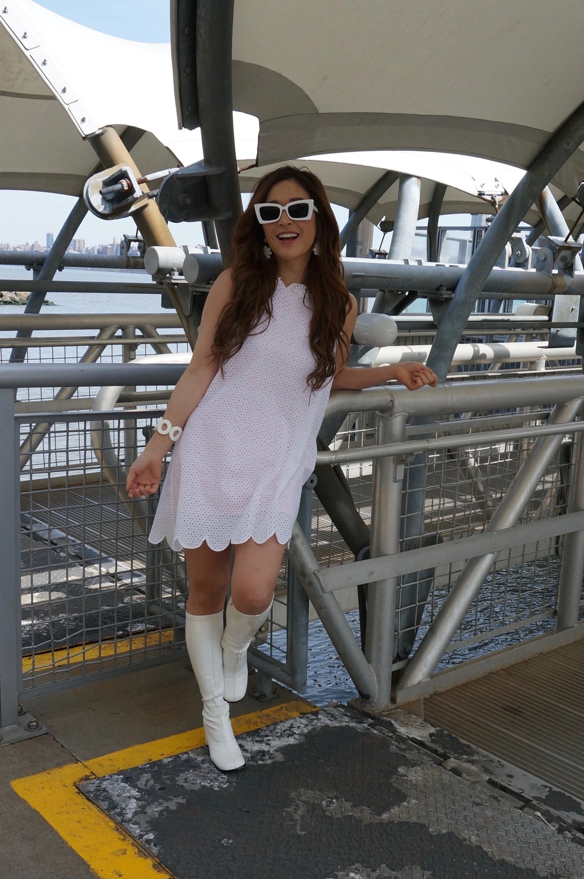 Model wearing an eyelet tent style dress with a peek through bubblegum lining, with a scalloped hem, and a daisy embroidered trim smiling at a ferry terminal.