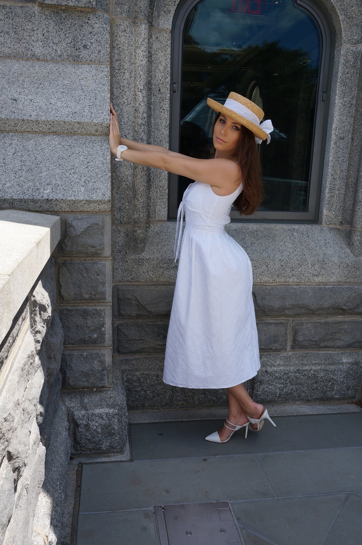 Model wearing a white floral eyelet fabric and a low V-neck with a bow top on a balcony in front of a window with her arms against the wall.