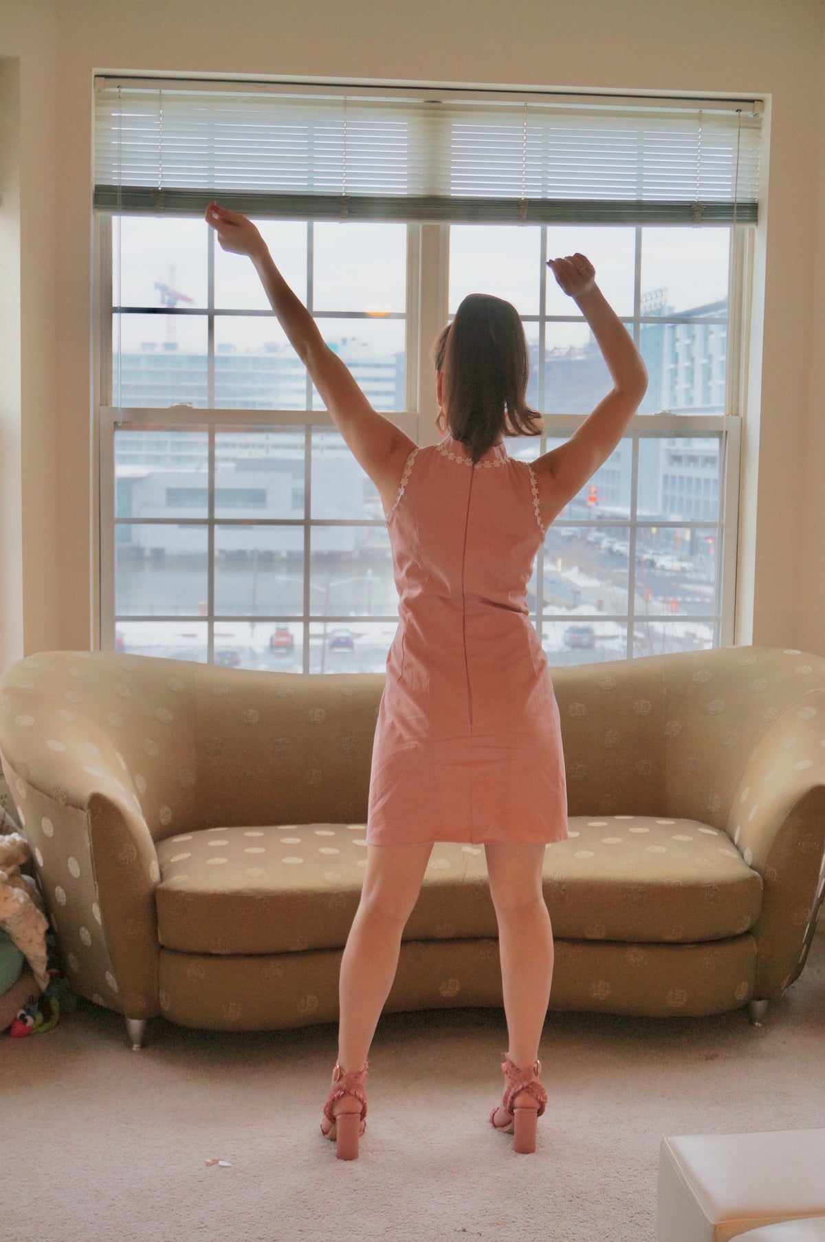 Model is wearing a pink dress with lace daisy trim and a front kick pleat looking off to the side in front of a brown couch with her arms up.