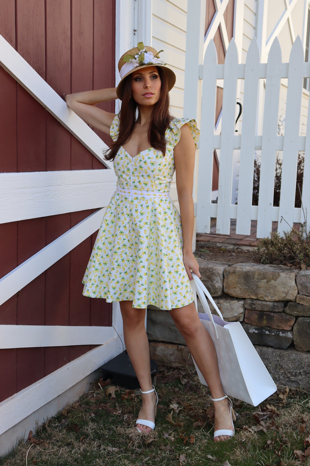 Model in lemon print dress  wearing a straw hat in front  of a barn, with left arm propped up against the barn looking off to the right.
