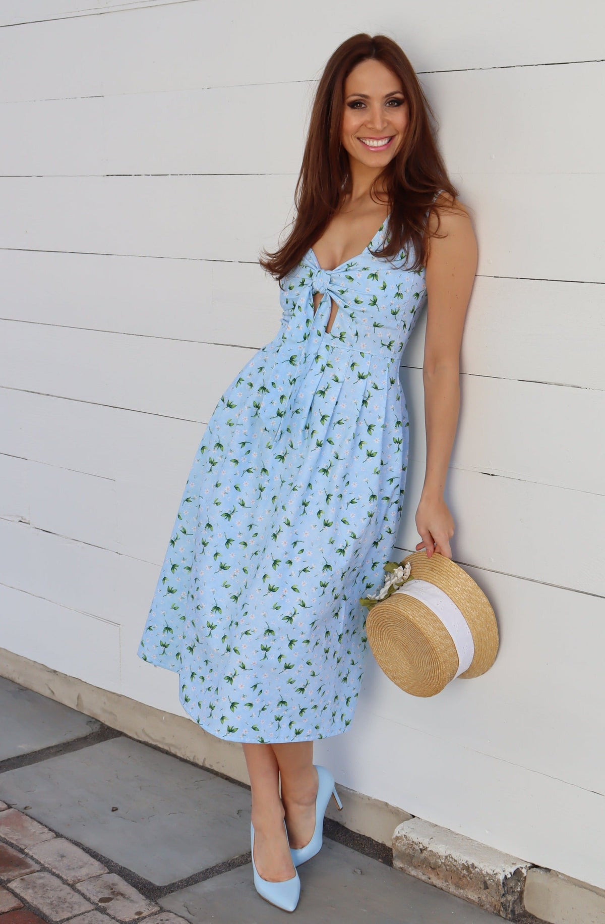 Model wearing a midi light blue dress with peek-a-boo bow front, in front of a white wood wall.