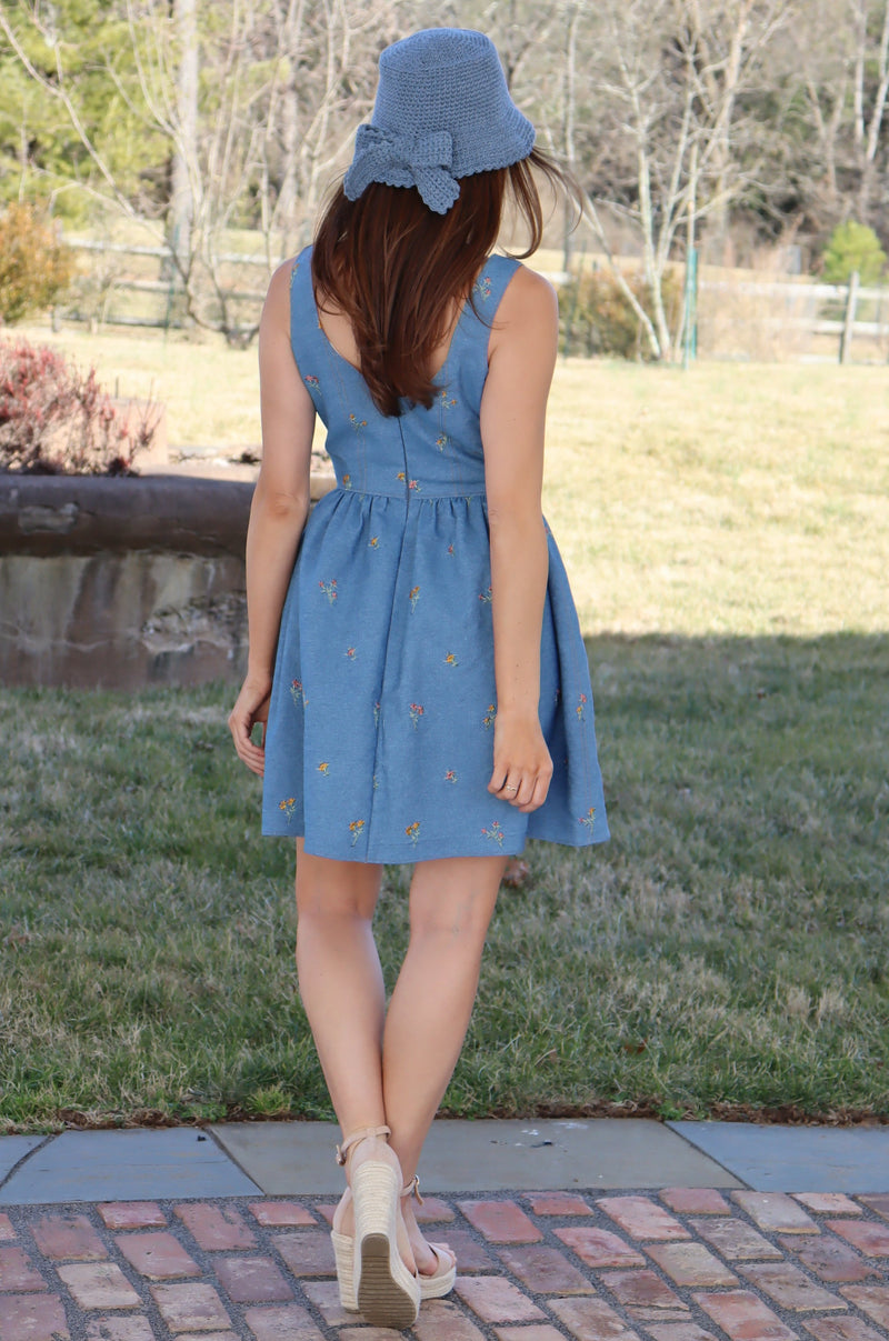 Back of model in floral embroidered denim dress wearing a hand crocheted blue bucket hat with a bow in front of a field of grass with left ankle crossing over the right ankle.