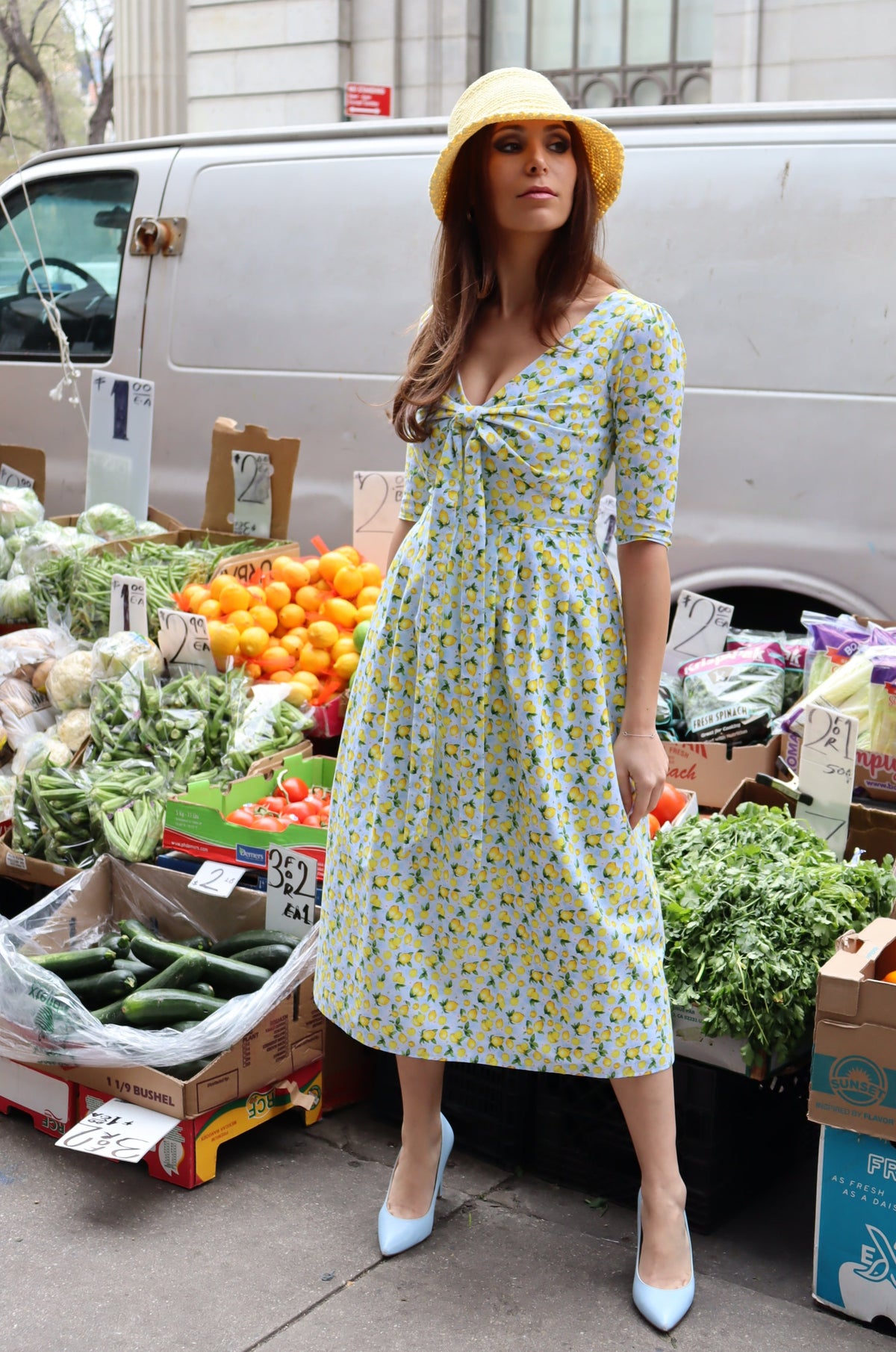 Model wearing a tan bucket hat and a midi dress in a lemon print of aqua blue and  yellow in front of a fruit display at a farmer's market.