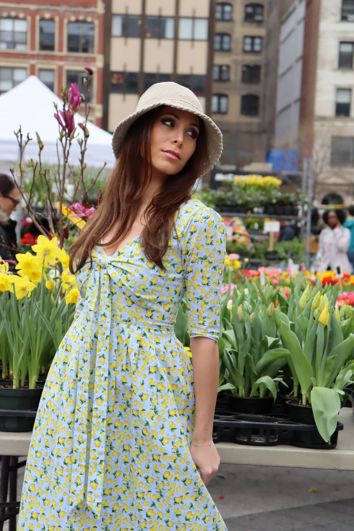 Close up view of Model wearing a tan bucket hat and a midi dress in a lemon print of aqua blue and  yellow in front of a flower display at a farmer's market.