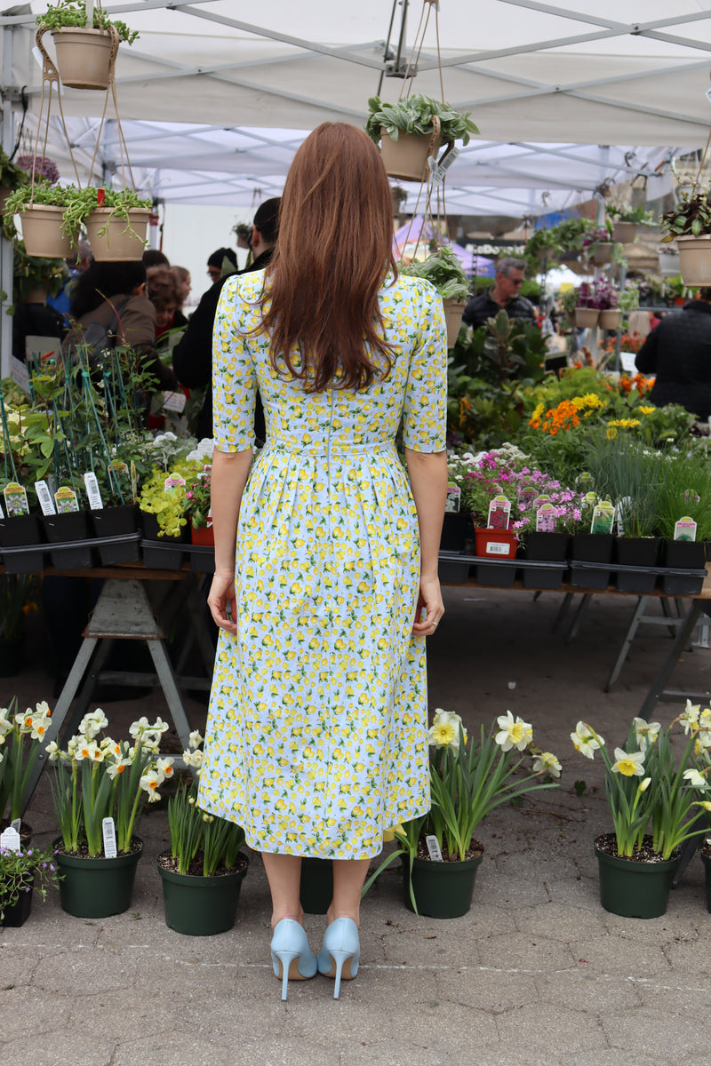 Back view of Model wearing a midi dress in a lemon print of aqua blue and  yellow in front of a flower display at a farmer's market.