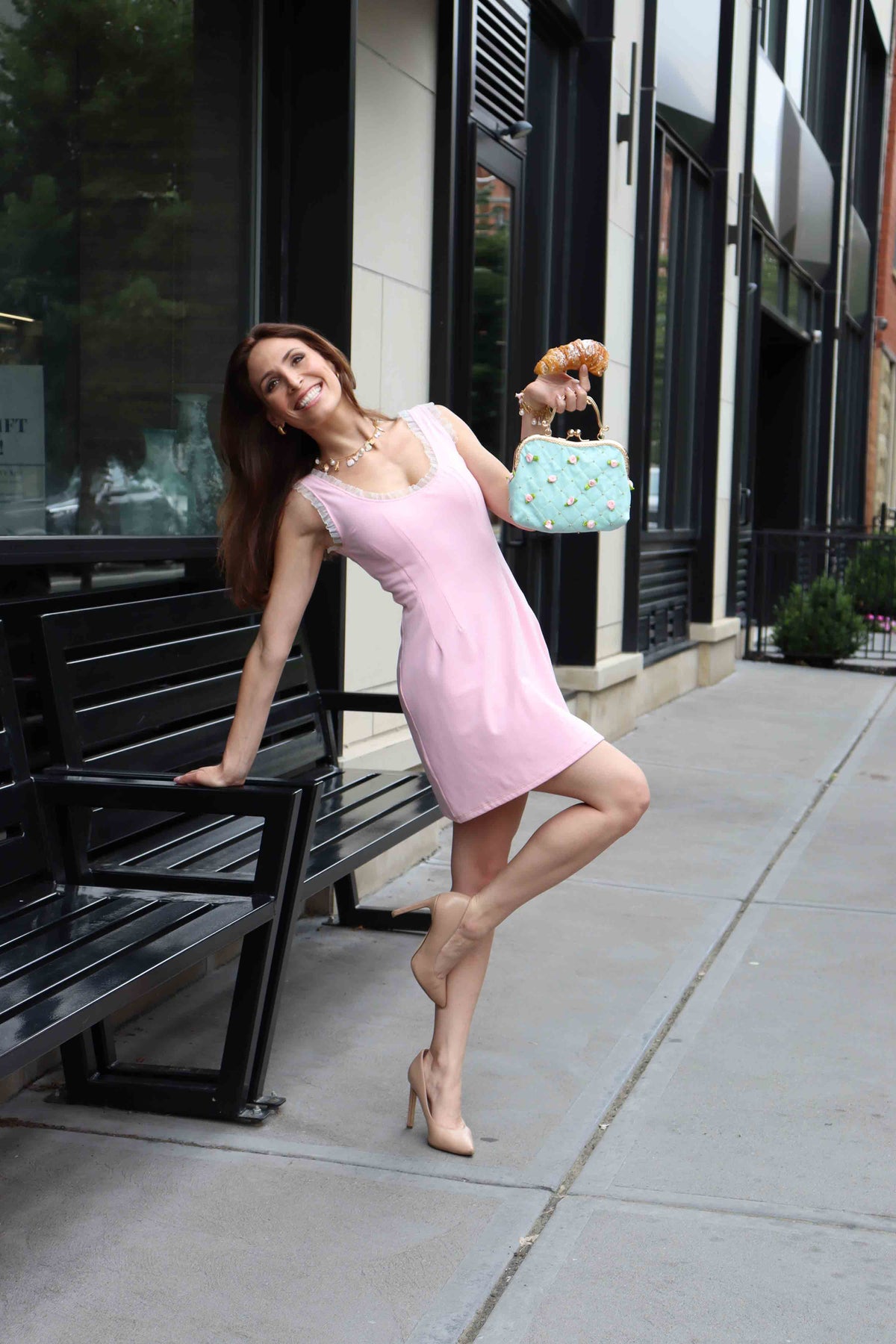 Back of model wearing pink silky stretch cotton dress leaning back in front of black bench smiling with one leg bent.