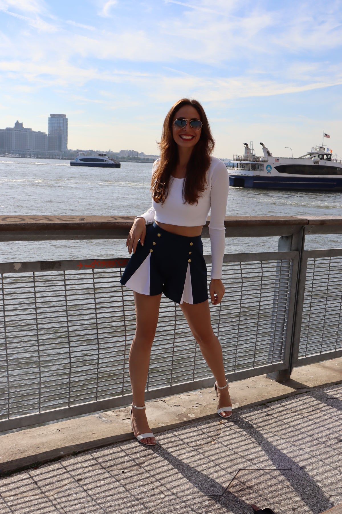 Model wearing scoop neck long sleeve white crop top and blue and white spinnaker shorts with gold buttons in front of the water with a hand on her hip smiling.