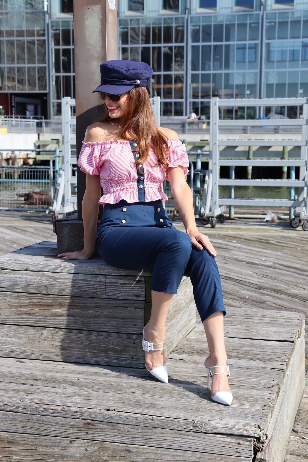 Model wearing jib top, navy cotton port capris and blue hat sitting down and smiling.