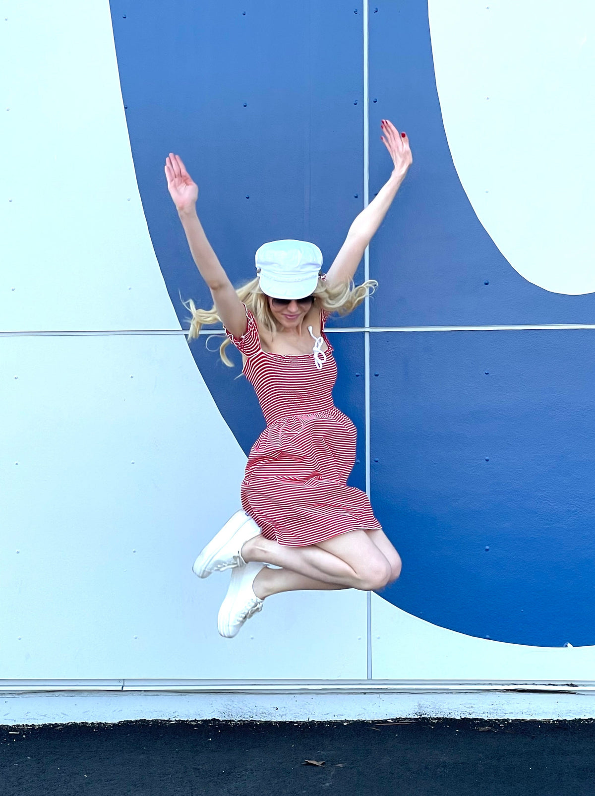 Model wearing red and white striped shirred dress and white hat jumping with both hands in the air.