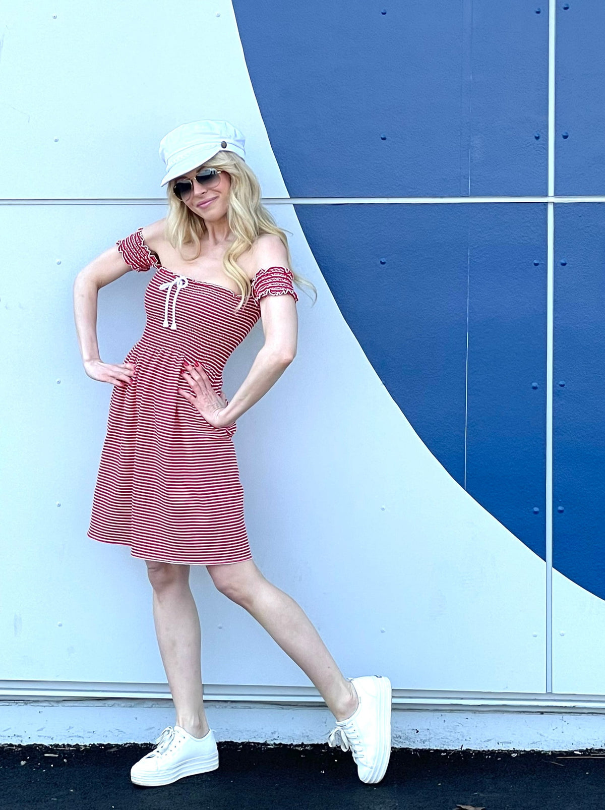 Model wearing red and white striped shirred dress and white hat with hands on her hips.