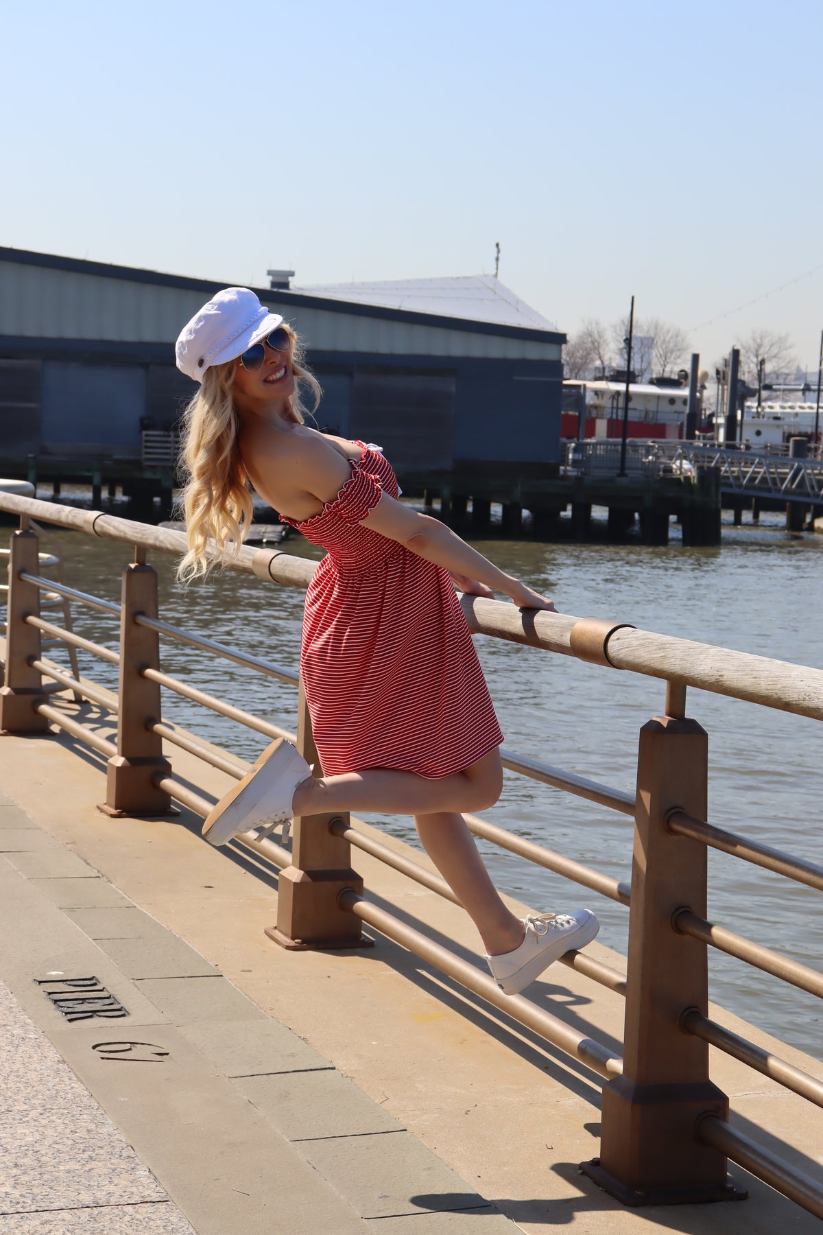 Model wearing red and white striped shirred dress and white hat leaning back from a railing smiling.