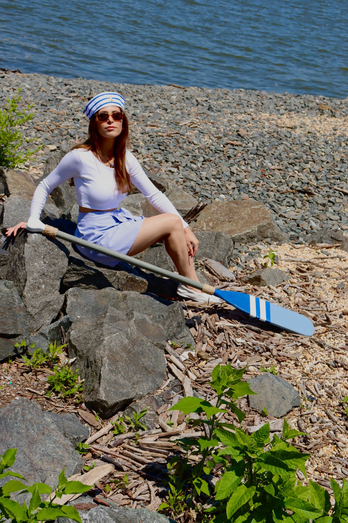 Model wearing a white and blue striped sailing hat, a white long sleeve crop top, and a light blue cotton print skirt with a bow, sitting down with an oar in front.