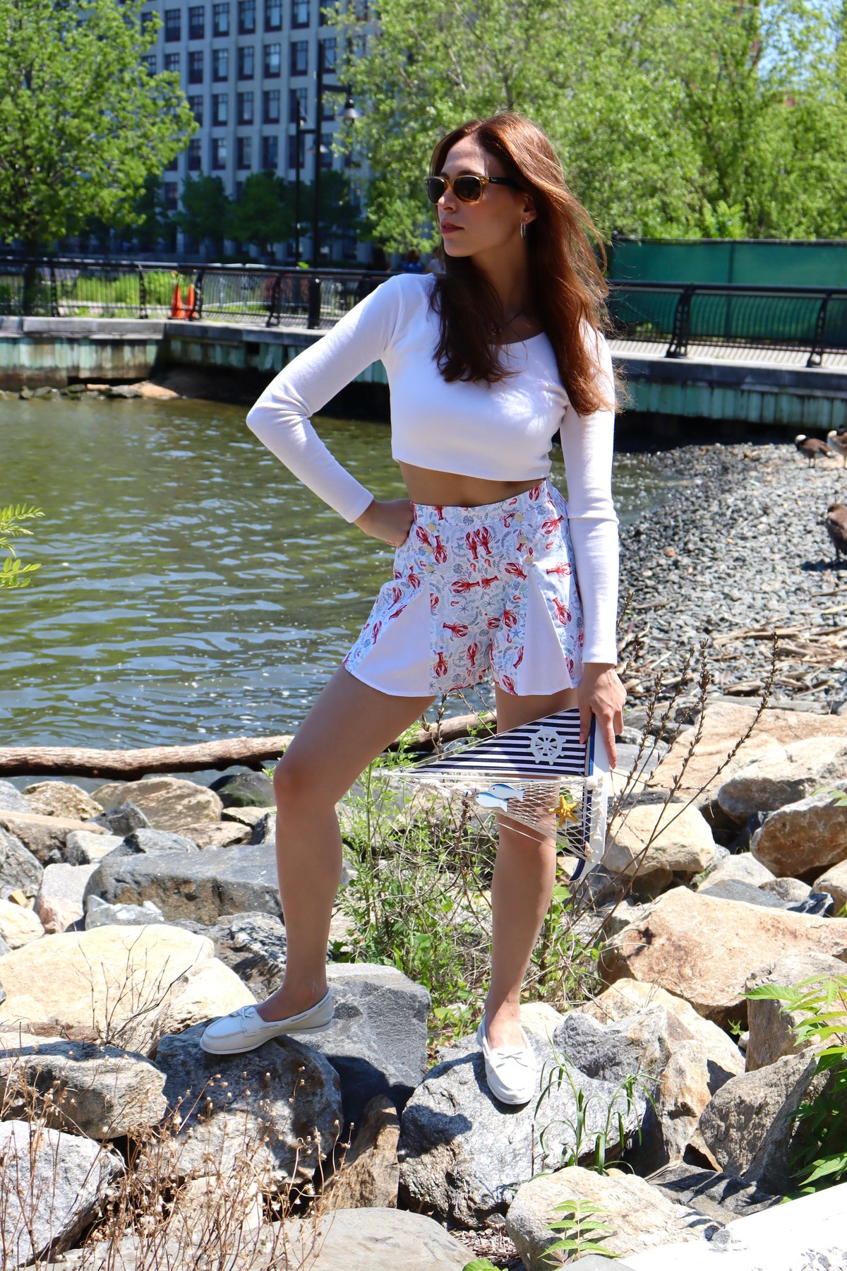 Model wearing pleated blue and red lobster print cotton shorts and white long sleeve crop top with her hand on her hip looking off to the side, standing on rocks in front of  a river.