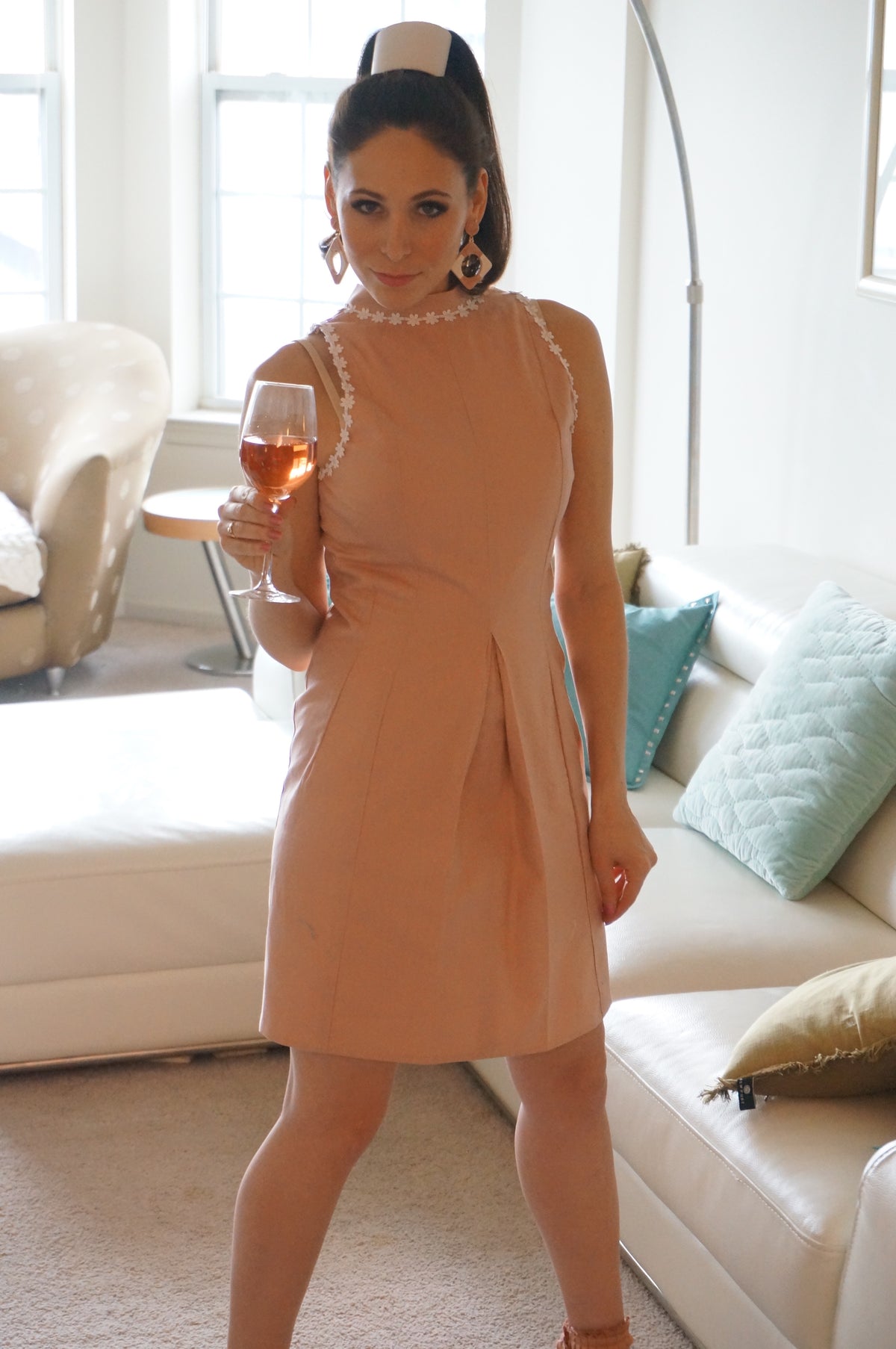 Model is wearing a pink dress with lace daisy trim and a front kick pleat looking off to the side in front of a couch holding a wineglass.