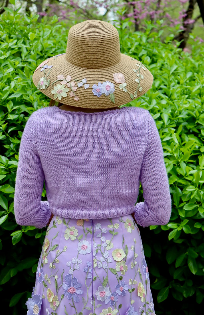 Model facing backwards to show back view of lilac cropped sweater with matching lilac appliquéd floral dress and floral detailed straw sun hat.