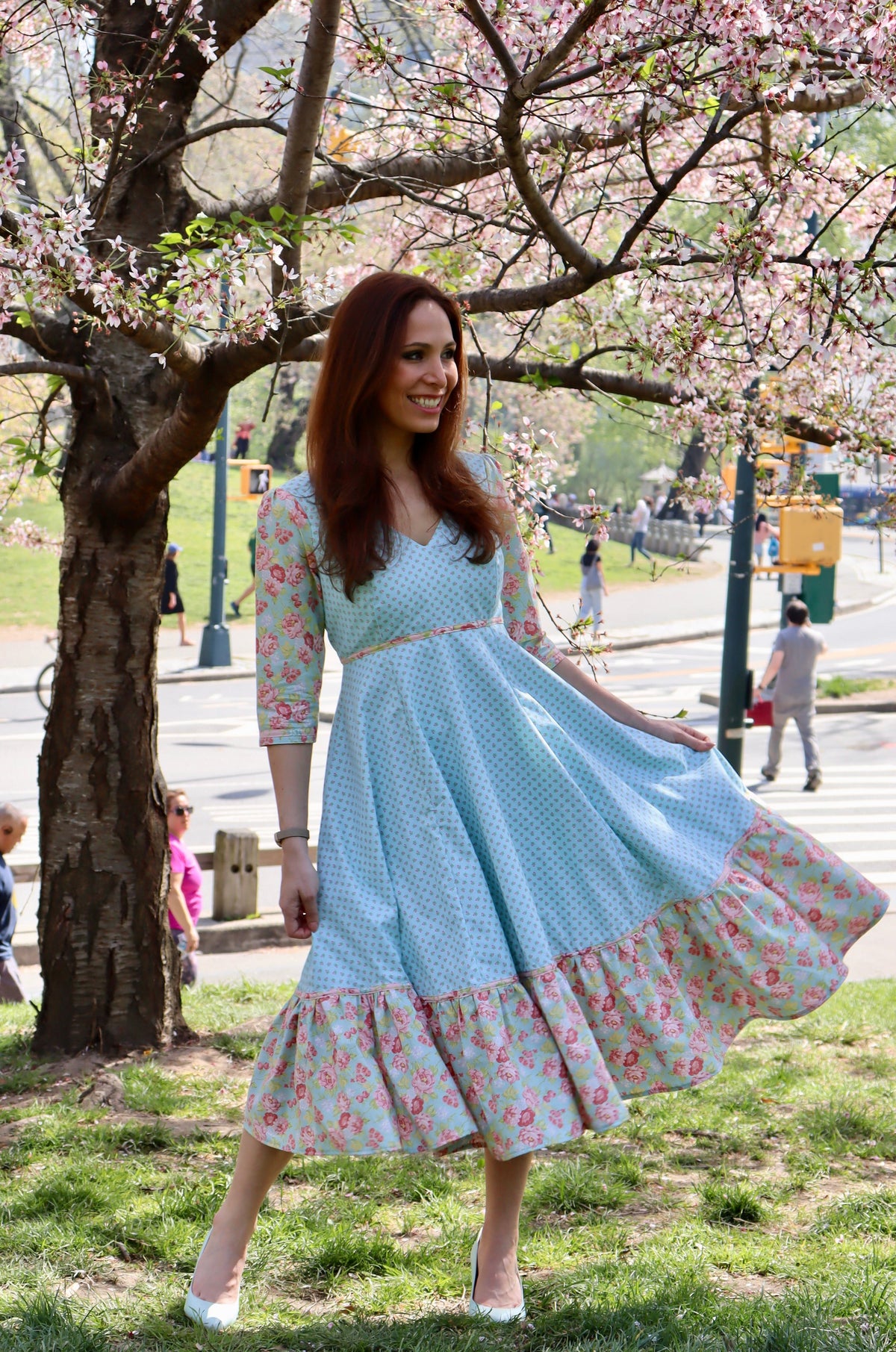Model in aqua blue print midi dress with sleeves and lower ruffle in print and blue rose print holding out skirt and standing  in front of a cherry tree.