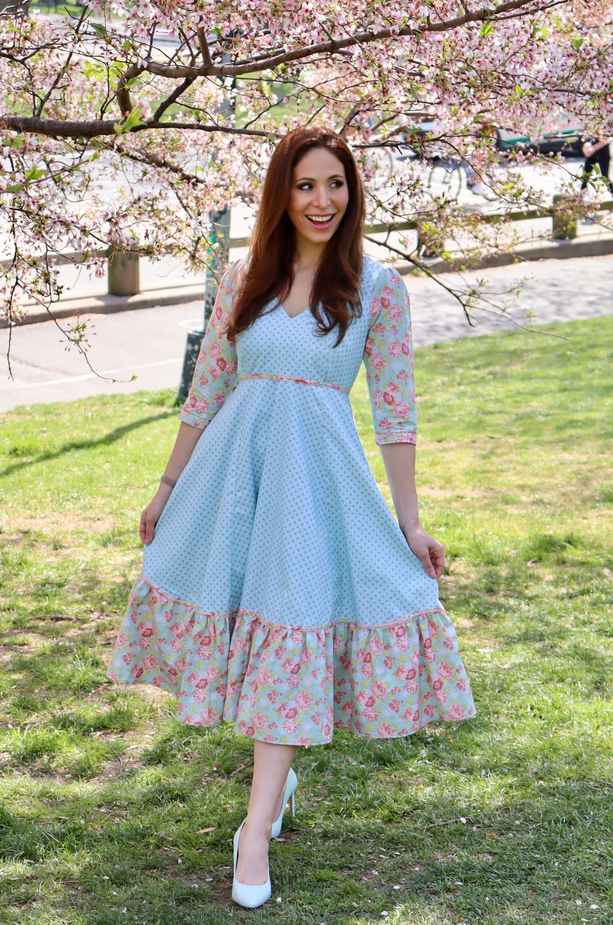 Model in aqua blue print midi dress with sleeves and lower ruffle in print and blue rose print holding out skirt and standing in front of a cherry tree.