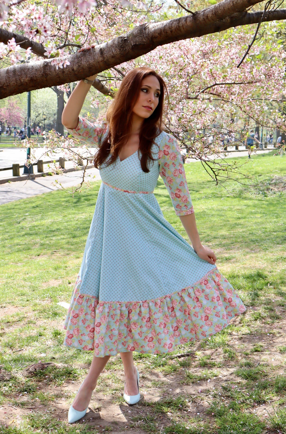 Model in aqua blue print midi dress with sleeves and lower ruffle in print and blue rose print holding out skirt and standing holding onto a tree limb.