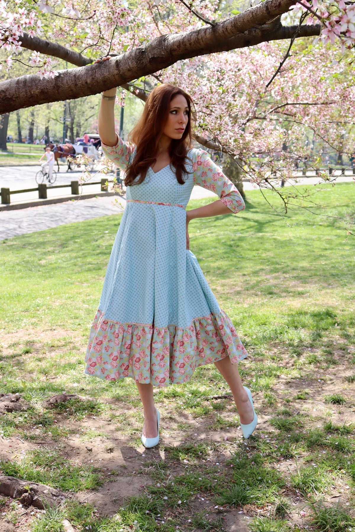 Model in aqua blue print midi dress with sleeves and lower ruffle in print and blue rose print standing holding onto a tree limb.