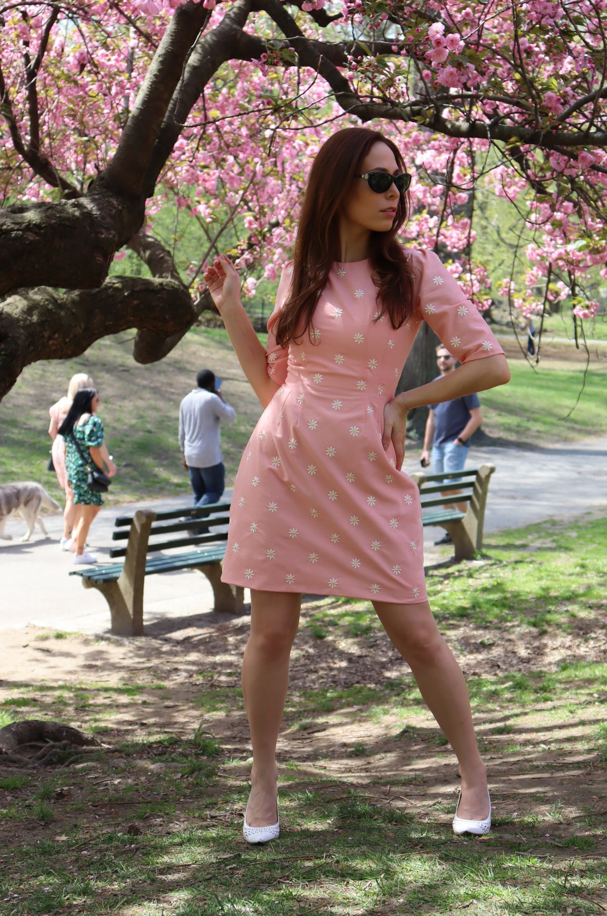 Model in peachy pink short dress with white daisy appliques looking to the side in front of a cherry tree.