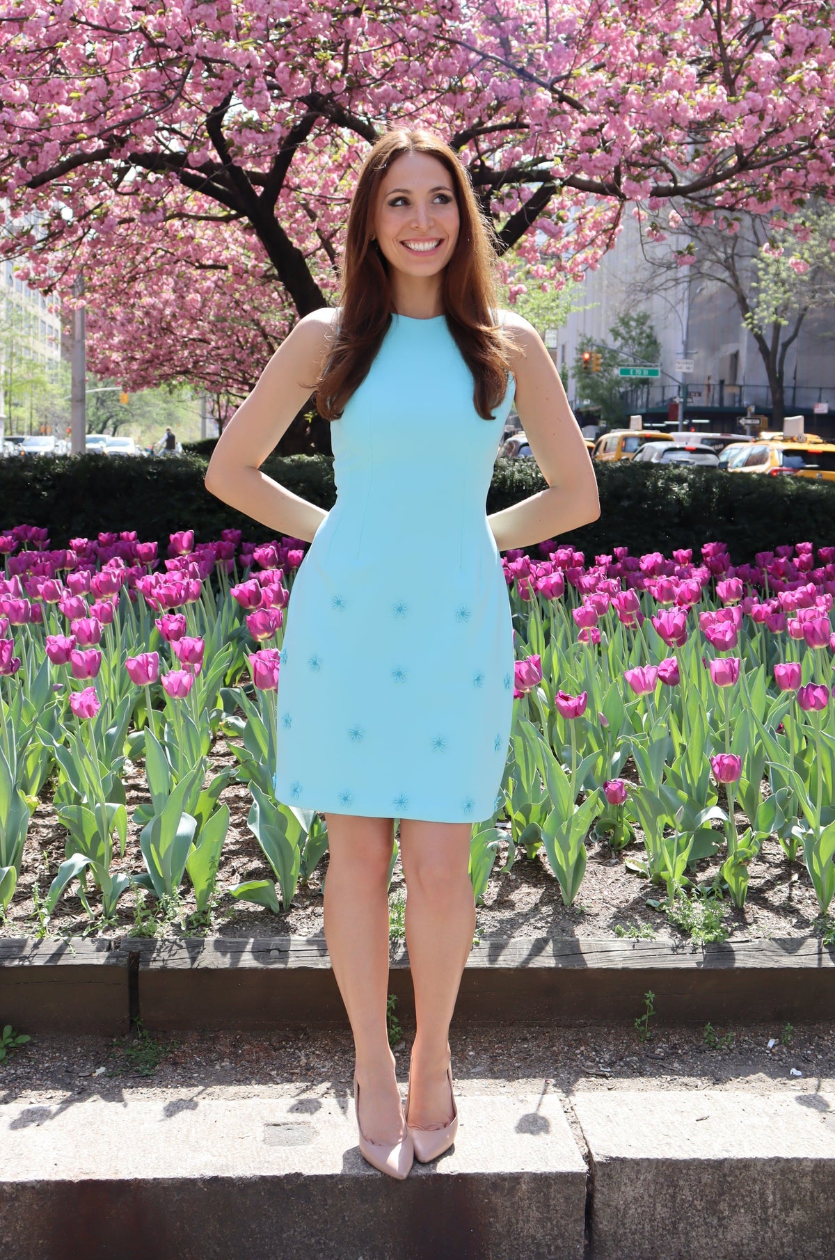 Model smiling and wearing a short aqua dress with aqua daisy appliques in front of purple tulips on Park Ave.