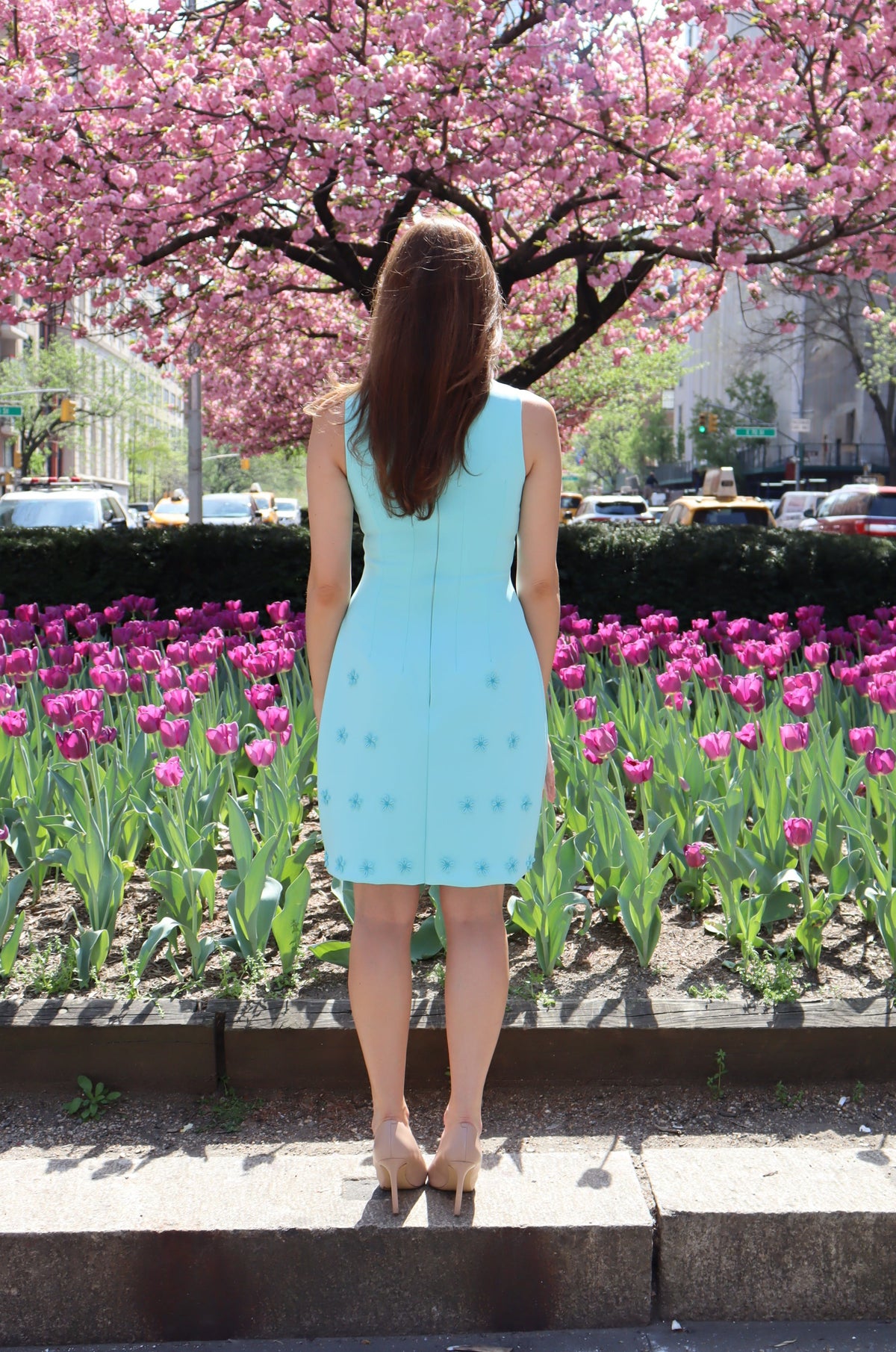 Back view of Model wearing a short aqua dress with aqua daisy appliques in front of purple tulips on Park Ave.