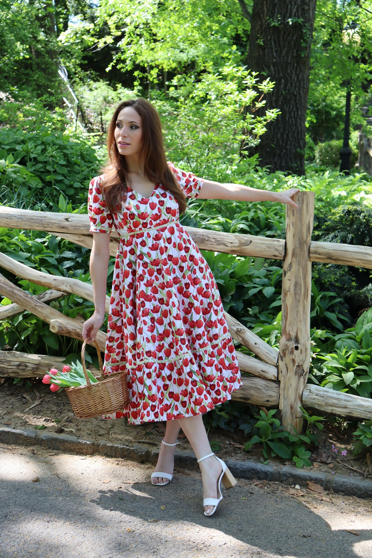 Model wearing midi length dress with a tulip print on white and holding a basket of tulips in front of a wood fence.