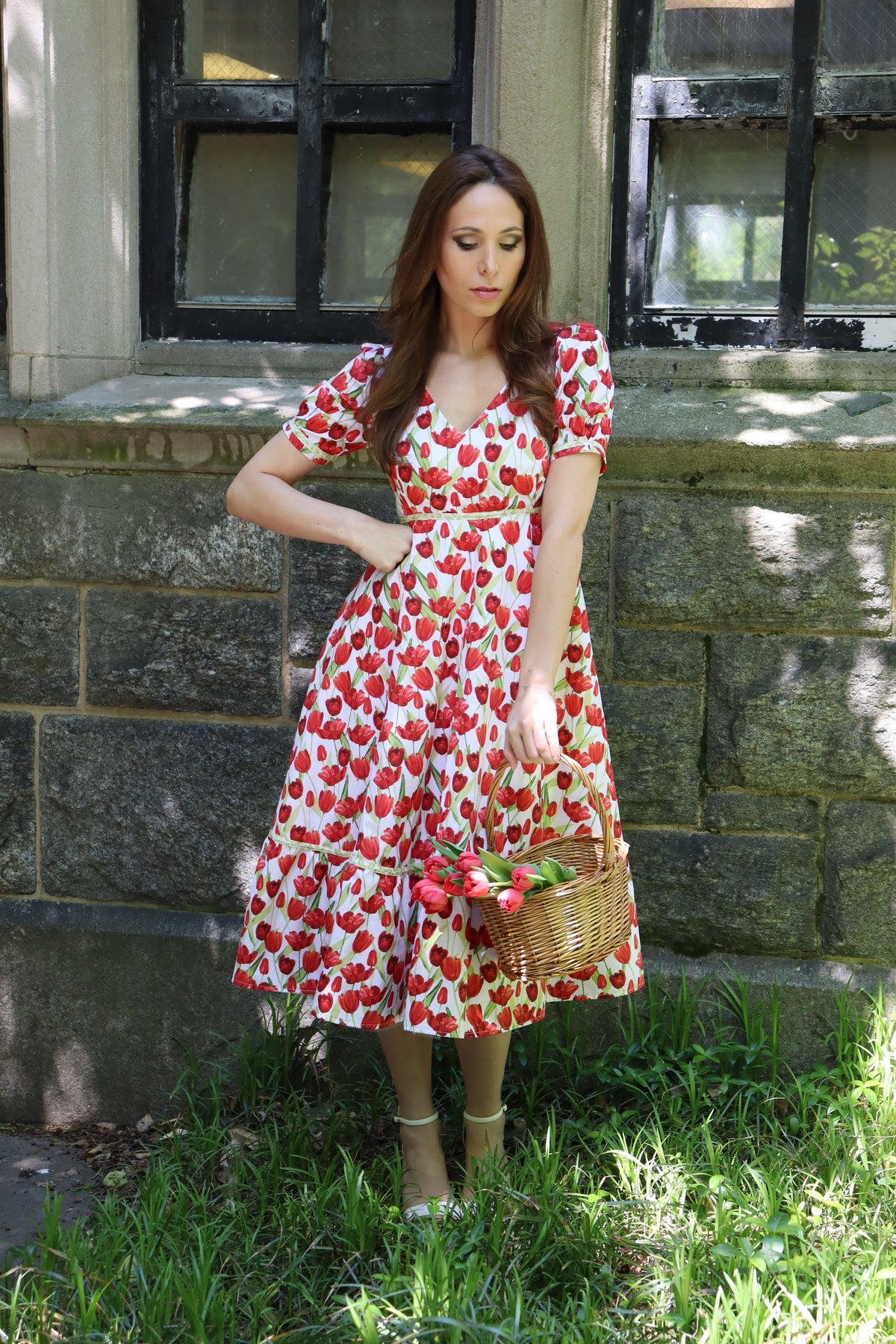 Model wearing midi length dress with a tulip print on white and holding a basket of tulips looking downward.