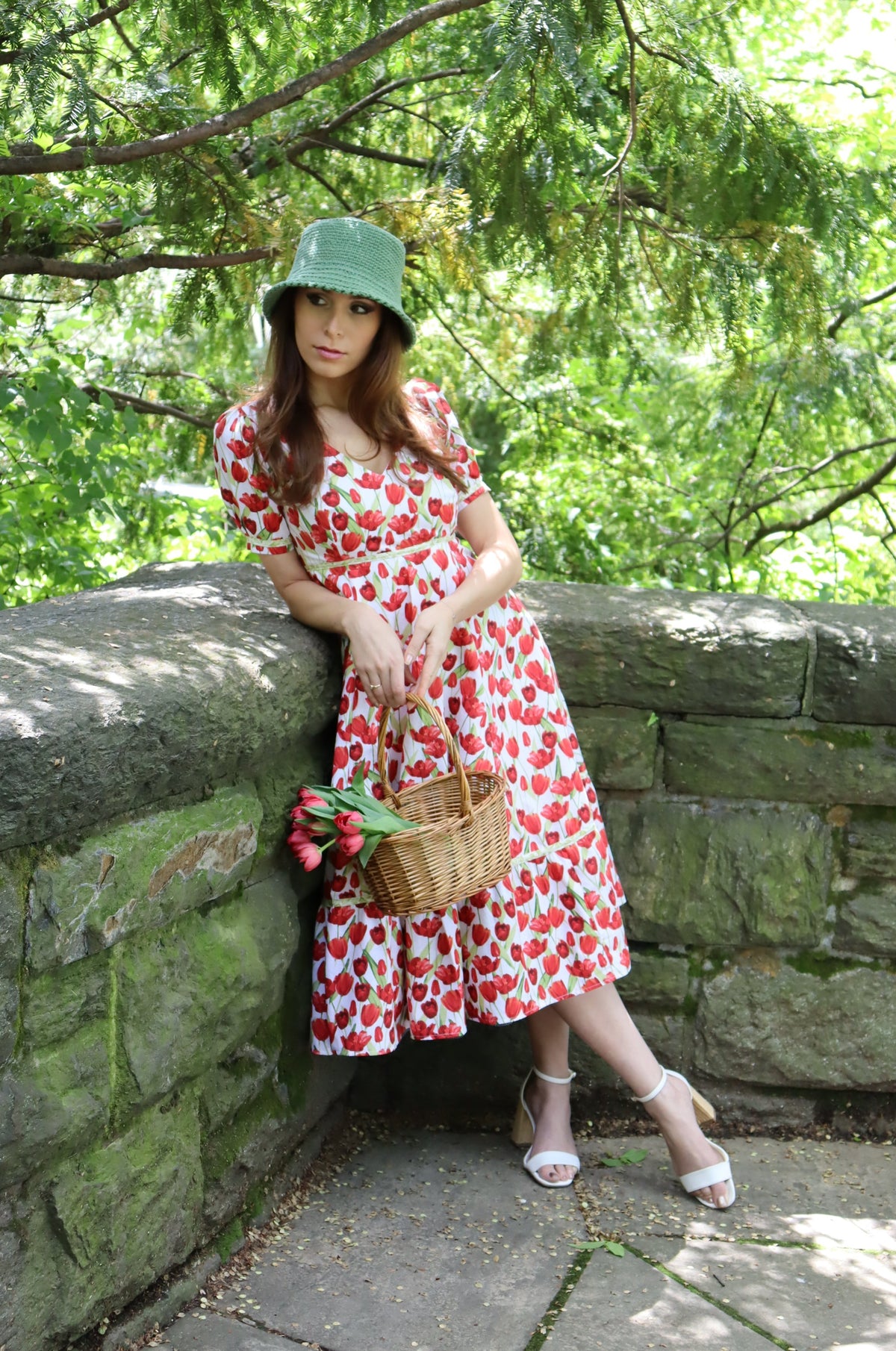 Model wearing midi length dress with a tulip print on white and wearing a green hat leaning on a stone wall.