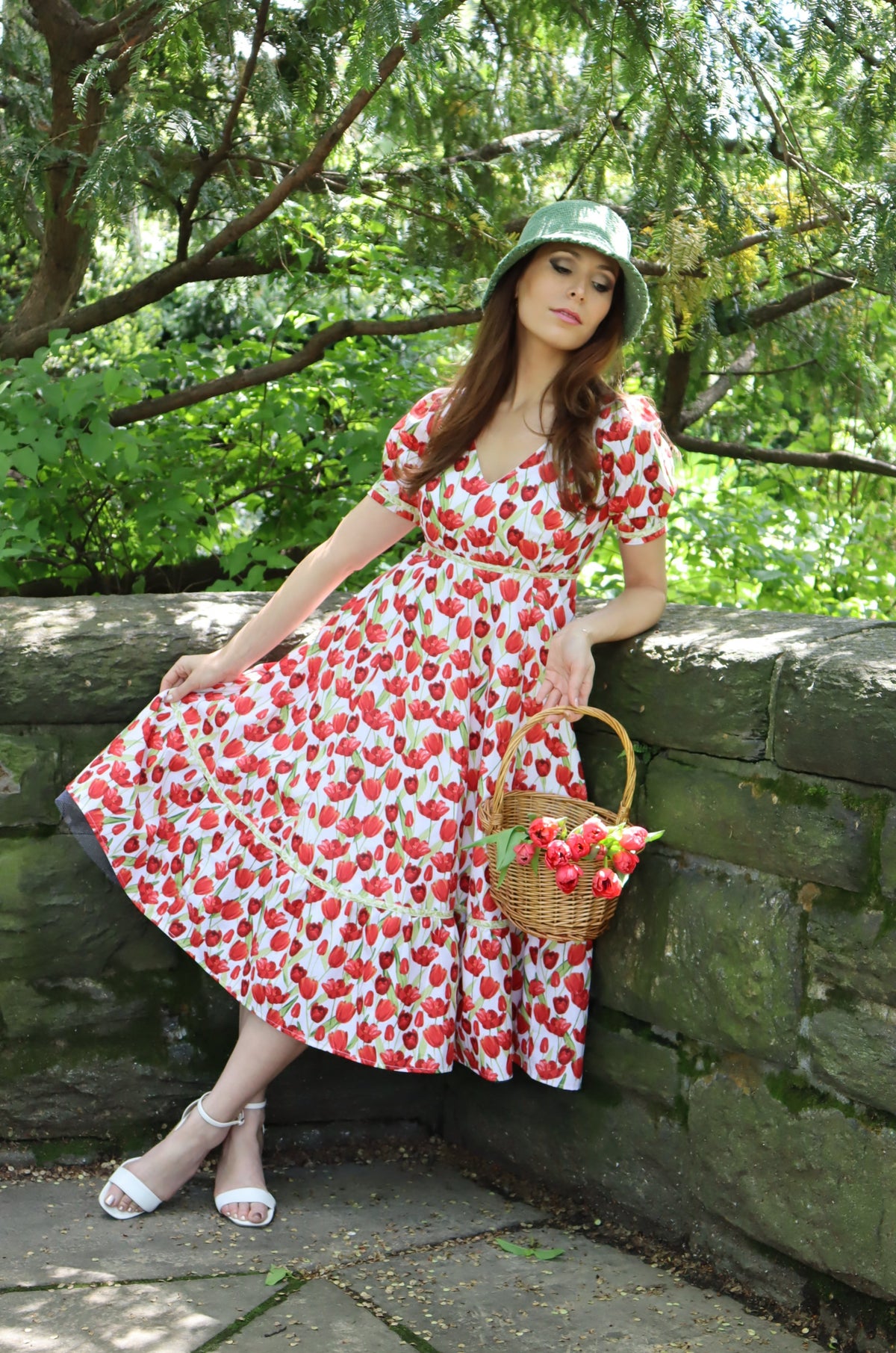 Model wearing midi length dress with a tulip print on white and wearing a green hat and holding a basket of tulips with her eyes downward.