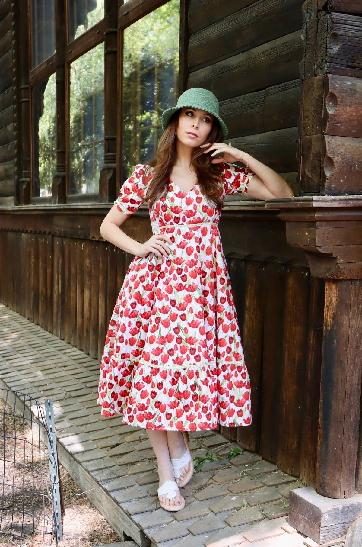 Model wearing midi length dress with a tulip print on white and wearing a green hat against a log cabin.