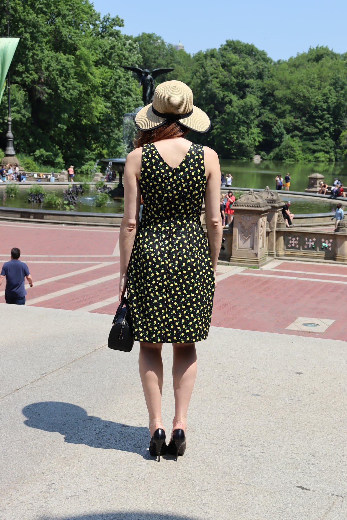 Back view of model in a black and yellow lemon print short dress wearing a sunhat with a black ribbon standing in Central Park before a fountain and a lake.balustrad.