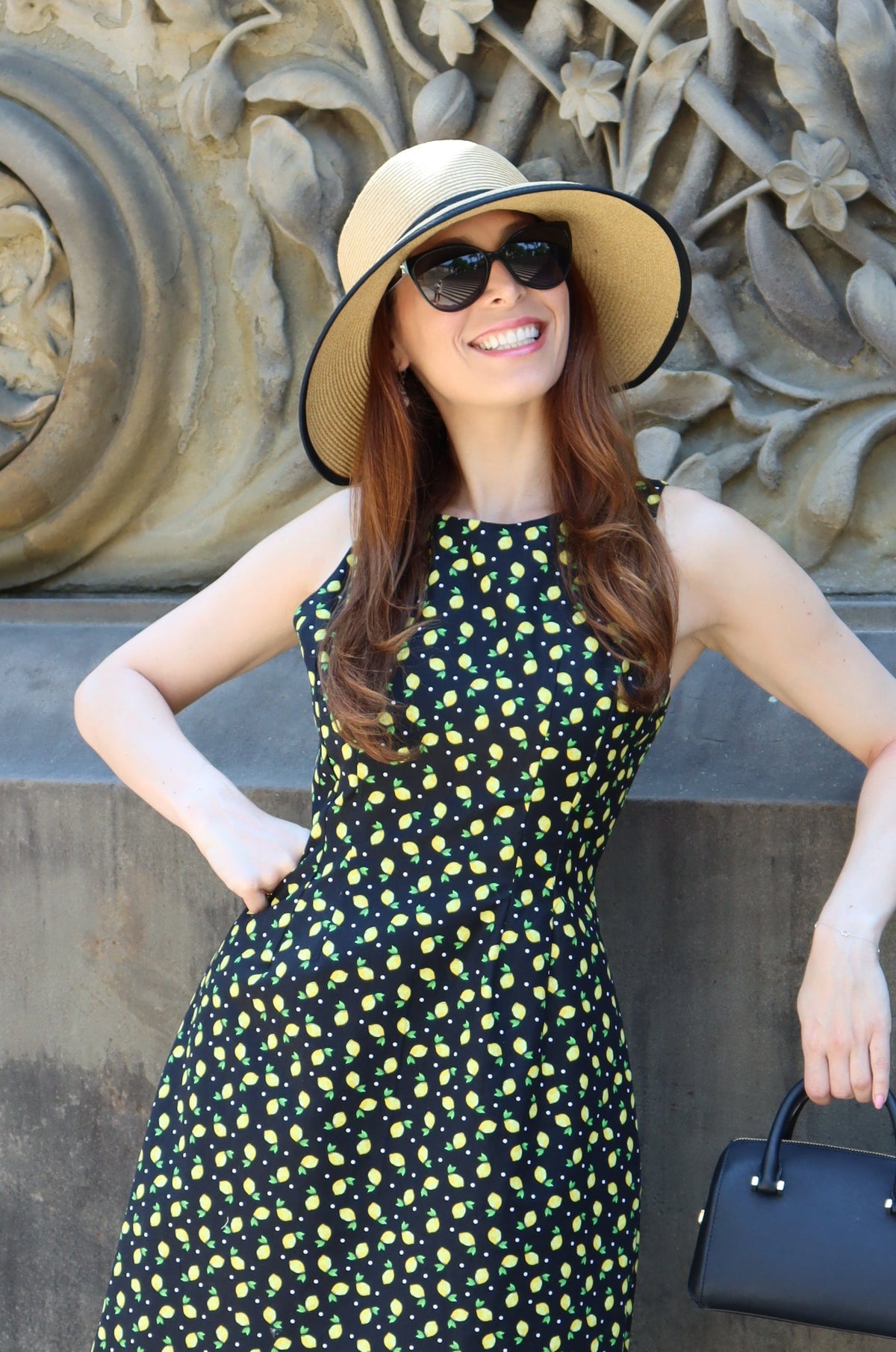 Close up photo of model in a black and yellow lemon print short dress wearing a sunhat with a black ribbon standing in front of a stone balustrad.