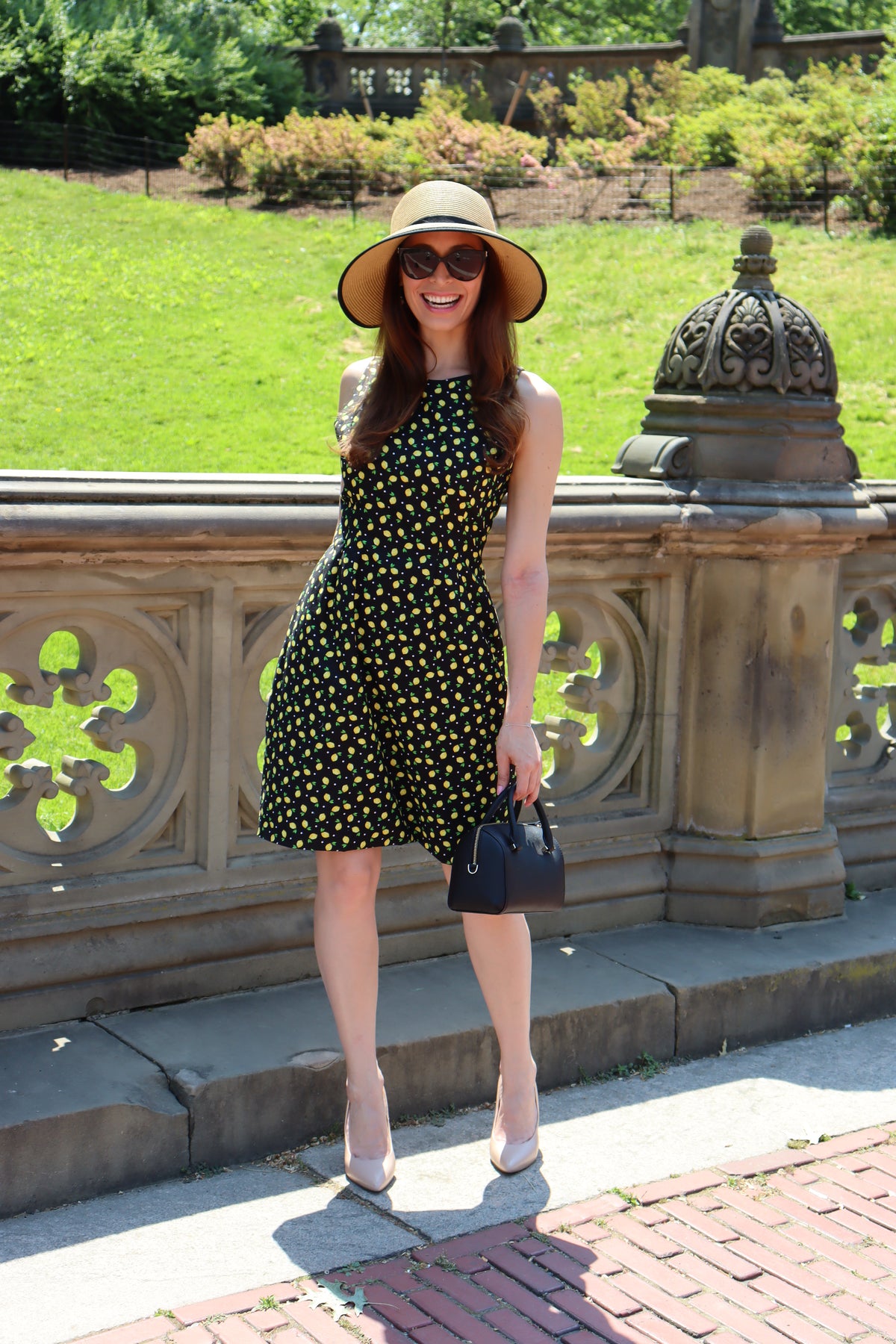 Model in a black and yellow lemon print short dress wearing a sunhat with a black ribbon standing in front of a stone balustrad.