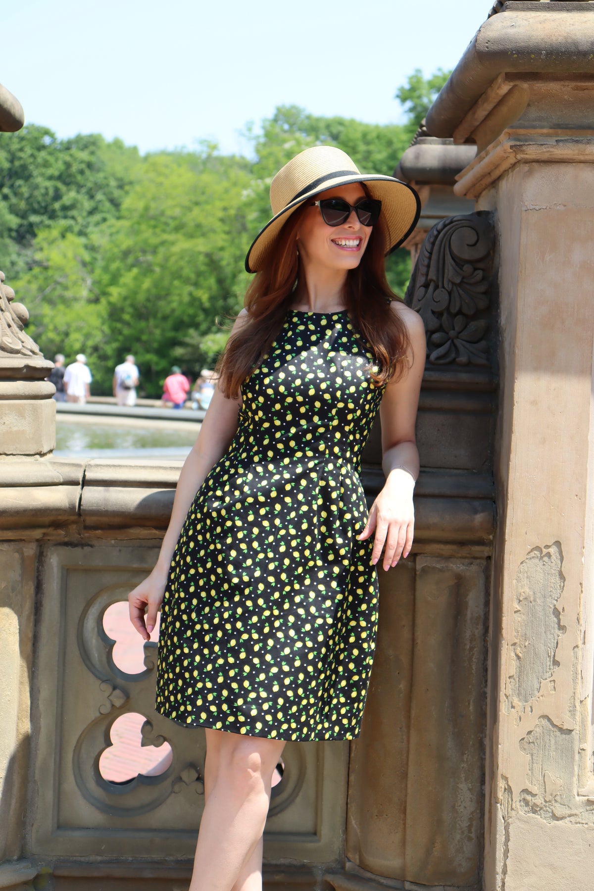 Model looking to the side in a black and yellow lemon print short dress wearing a sunhat with a black ribbon standing in front of a stone balustrad.
