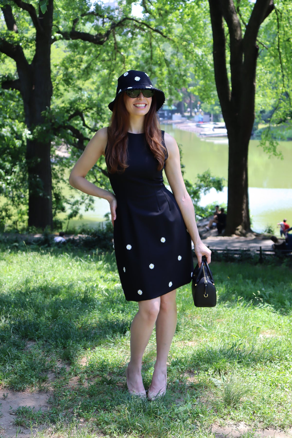 Model in a black dress with white daisy appliques standing in front of  trees and a lake.