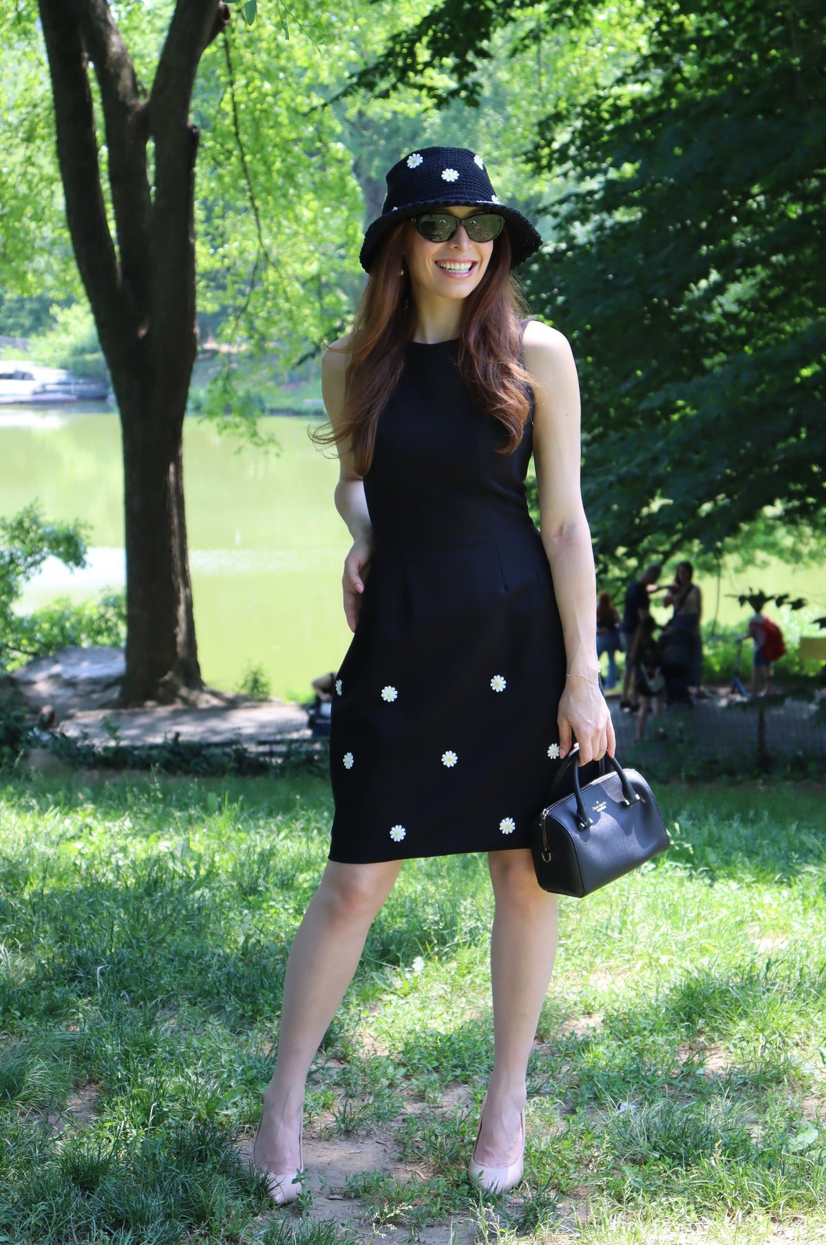 Model in a black dress with white daisy appliques standing with hand on hip  in front of a tree and a lake.