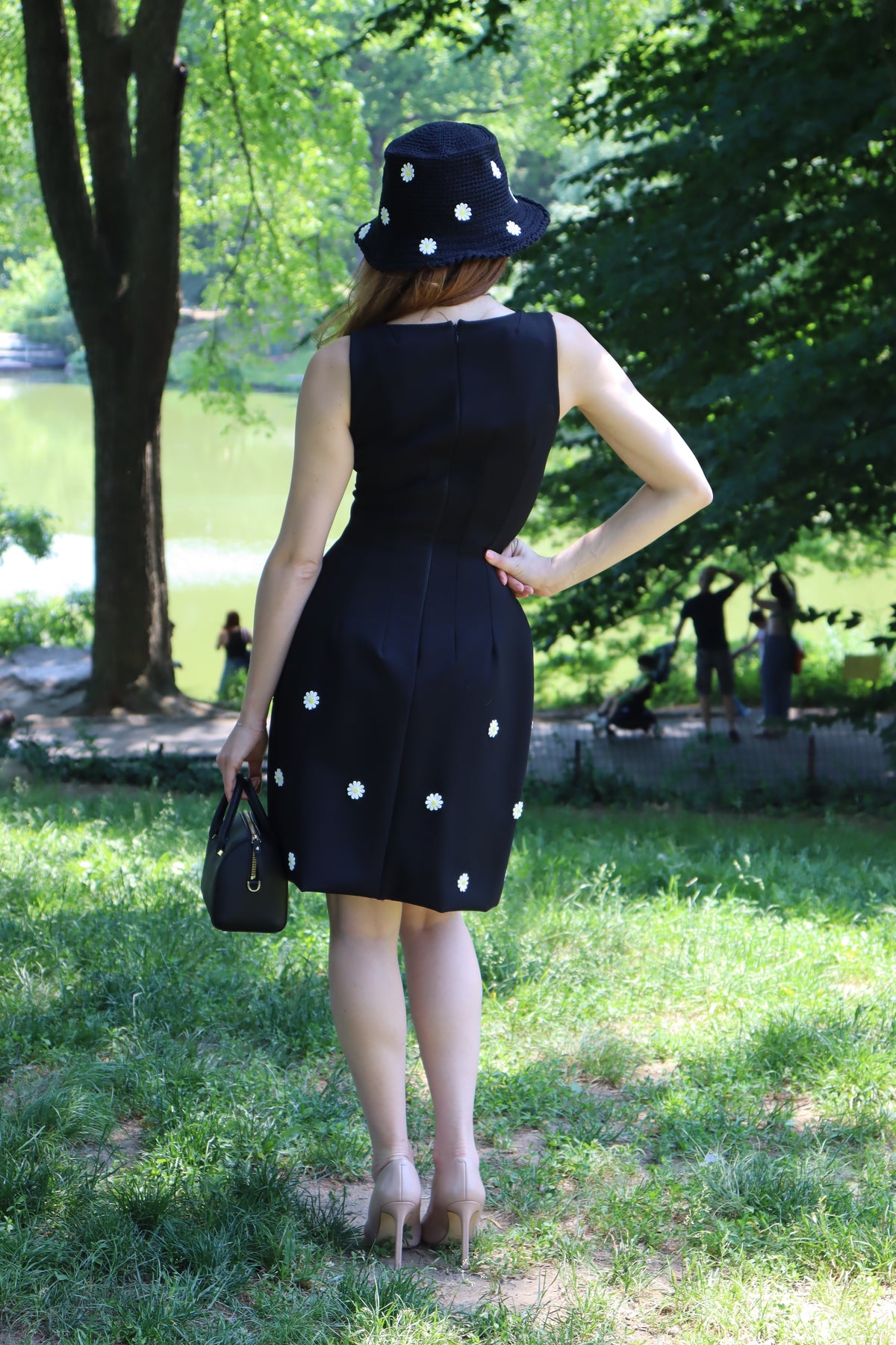 Back view of model in a black dress with white daisy appliques standing in front of a tree and a lake.