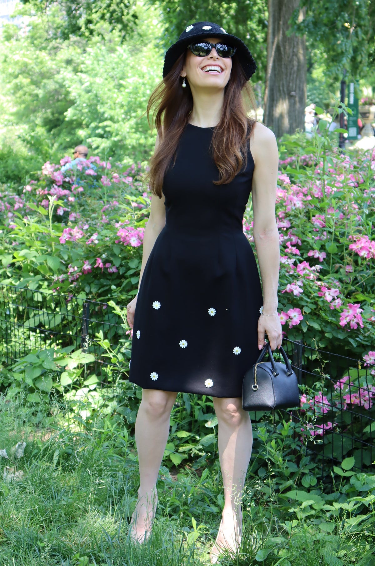 Model in a black dress with white daisy appliques and matching hat looking up standing in front of pink flowers.