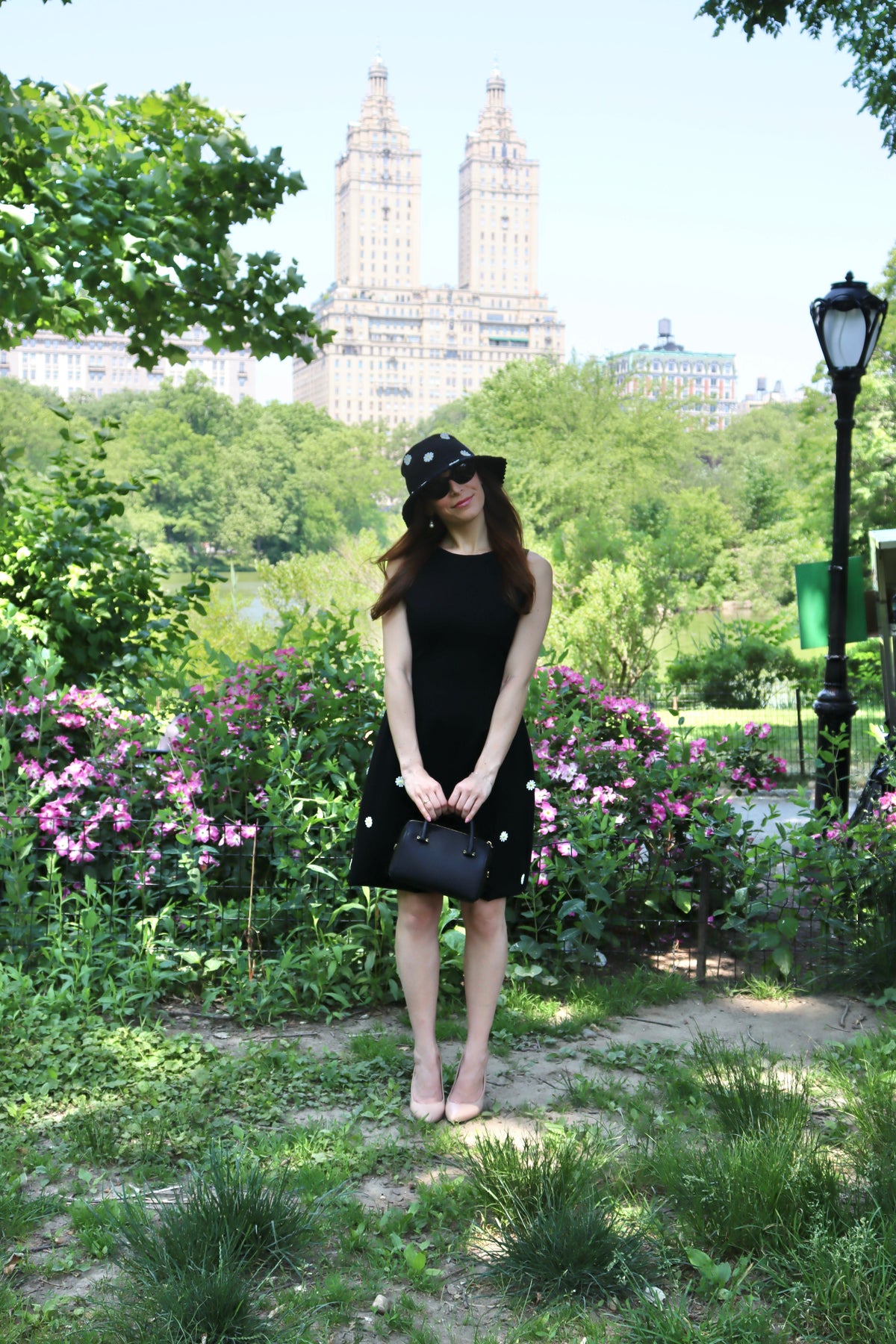 Model in a black dress with white daisy appliques and matching hat standing in front of pink flowers and a building.