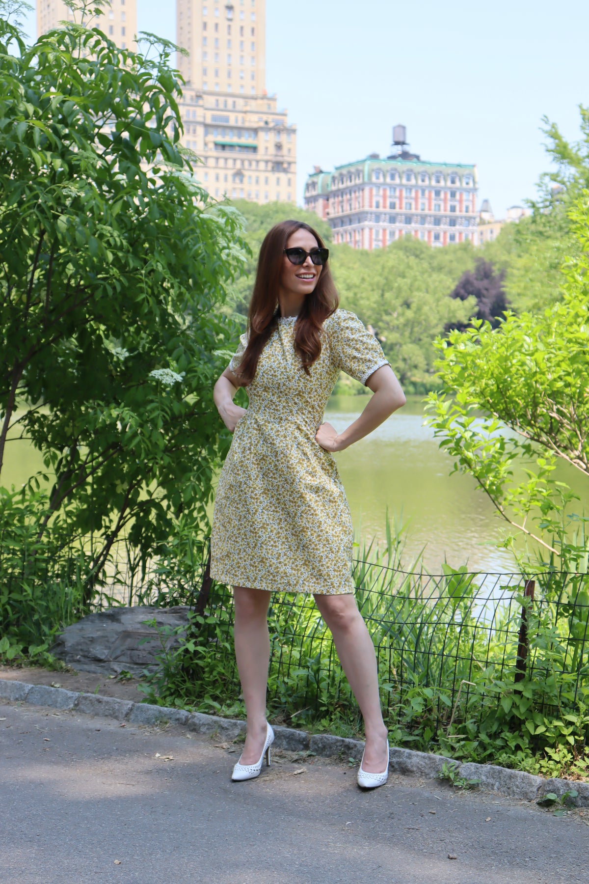 Photo of a model in short yellow floral dress with short sleeves and white daisy trim leaning standing in front of a lake with buildings in the background in Central Park.on a tree.