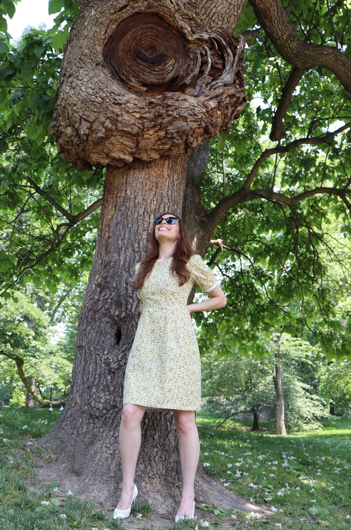 Model in short yellow floral dress with short sleeves and white daisy trim leaning on a tree.